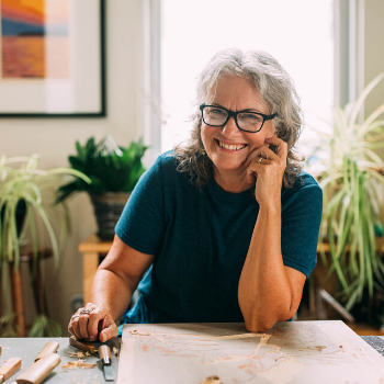 Mary sits at her studio desk. She is wearing a teal shirt and smiling.